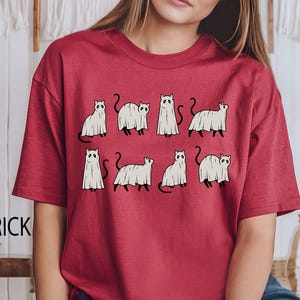 May include: Red t-shirt with a graphic of white ghost cats. The cats are in various poses, some with their tails up and some with their tails down.