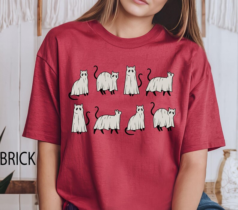 May include: Red t-shirt with a graphic of white ghost cats. The cats are in various poses, some with their tails up and some with their tails down.