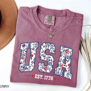 May include: Berry-colored t-shirt with the word "USA" in a floral design. The shirt also features the text "EST. 1776" and a small tag that says "Comfort Colors". A silver necklace with a pendant is draped across the shirt.
