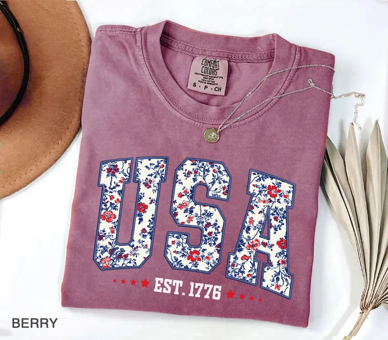 May include: Berry-colored t-shirt with the word "USA" in a floral design. The shirt also features the text "EST. 1776" and a small tag that says "Comfort Colors". A silver necklace with a pendant is draped across the shirt.