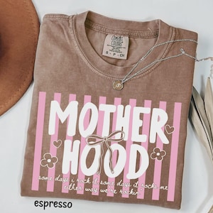 May include: Espresso-colored t-shirt with the word "MOTHERHOOD" in white, accented by pink stripes, hearts, and flowers. The shirt also features the text "some days i rock it some days it rocks me either way we're rockin'". A silver necklace is draped across the shirt.