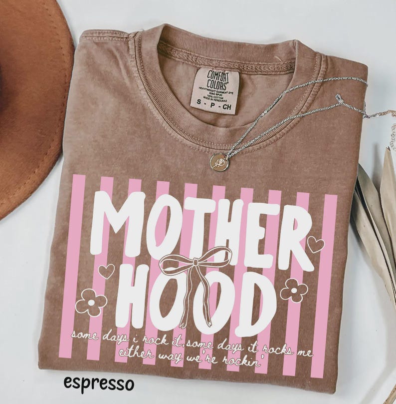 May include: Espresso-colored t-shirt with the word "MOTHERHOOD" in white, accented by pink stripes, hearts, and flowers. The shirt also features the text "some days i rock it some days it rocks me either way we're rockin'". A silver necklace is draped across the shirt.