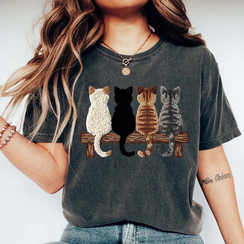 May include: Dark grey t-shirt with an embroidered design of four cats on a wooden bench. The cats are white, black, brown tabby, and grey tabby. The shirt has a vintage, worn appearance.