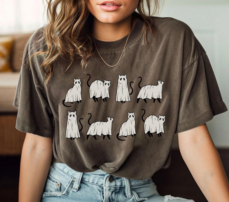 May include: A dark brown t-shirt with a graphic print of nine white ghost cats. The cats are in various poses, some are sitting, some are standing, and some are walking.