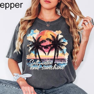 Op de afbeelding: Een grijs T-shirt met een afbeelding van palmbomen en een zonsondergang. De tekst "Salt water and sunshine" is op het T-shirt gedrukt.