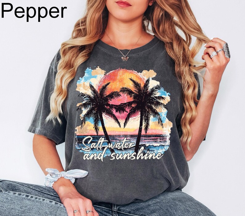 Op de afbeelding: Een grijs T-shirt met een afbeelding van palmbomen en een zonsondergang. De tekst "Salt water and sunshine" is op het T-shirt gedrukt.