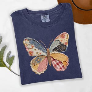May include: A navy blue t-shirt featuring a patchwork butterfly design. The butterfly wings are made of various patterned fabrics in shades of blue, pink, yellow, and red. The shirt has a crew neck and short sleeves.