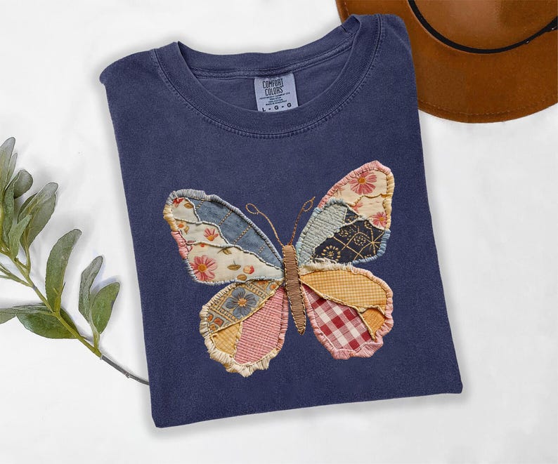 May include: A navy blue t-shirt featuring a patchwork butterfly design. The butterfly wings are made of various patterned fabrics in shades of blue, pink, yellow, and red. The shirt has a crew neck and short sleeves.