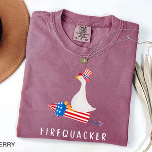 K&ouml;nnte beinhalten: Ein beerenfarbenes Kurzarm-T-Shirt mit einer Grafik einer wei&szlig;en Ente, die einen rot-wei&szlig;-blau gestreiften Zylinder tr&auml;gt und auf einer Feuerwerksrakete mit sternenbesetztem blauem Band reitet. Der Text FIREQUACKER ist darunter in Wei&szlig; gedruckt.