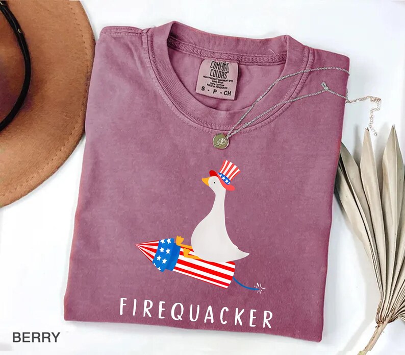 K&ouml;nnte beinhalten: Ein beerenfarbenes Kurzarm-T-Shirt mit einer Grafik einer wei&szlig;en Ente, die einen rot-wei&szlig;-blau gestreiften Zylinder tr&auml;gt und auf einer Feuerwerksrakete mit sternenbesetztem blauem Band reitet. Der Text FIREQUACKER ist darunter in Wei&szlig; gedruckt.