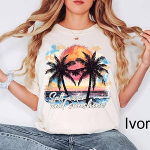 Op de afbeelding: Een wit T-shirt met een afbeelding van palmbomen en een zonsondergang. De tekst "Salt water and sunshine" is onder de afbeelding gedrukt. Het shirt wordt gedragen door een model met lang blond haar. De kleur van het shirt is ivoor.