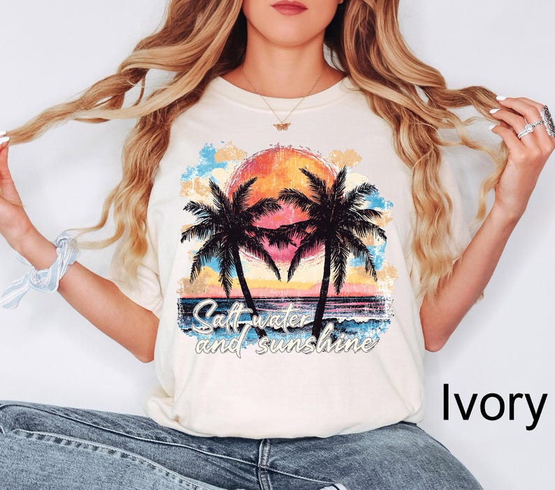 Op de afbeelding: Een wit T-shirt met een afbeelding van palmbomen en een zonsondergang. De tekst "Salt water and sunshine" is onder de afbeelding gedrukt. Het shirt wordt gedragen door een model met lang blond haar. De kleur van het shirt is ivoor.