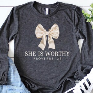 May include: A charcoal grey long-sleeved t-shirt with a large beige bow and the text "She is Worthy Proverbs 31" printed in white.