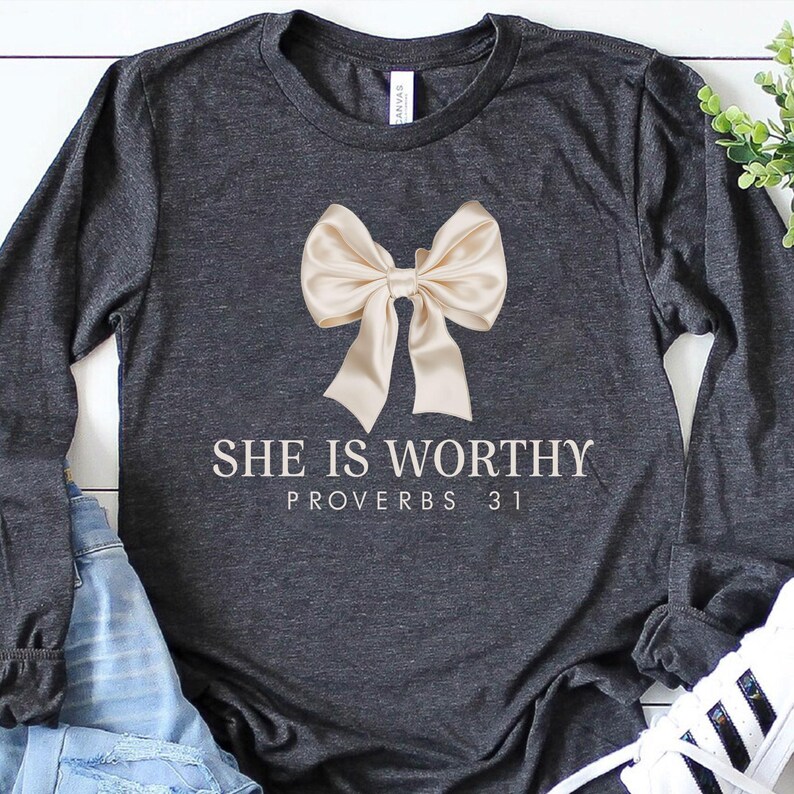 May include: A charcoal grey long-sleeved t-shirt with a large beige bow and the text "She is Worthy Proverbs 31" printed in white.