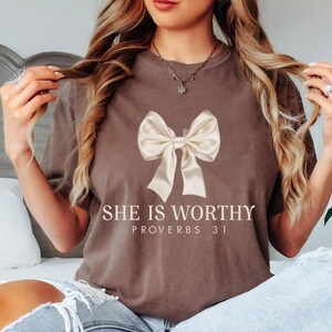 May include: A brown t-shirt with a white bow and the text "She is Worthy Proverbs 31" printed on the front.