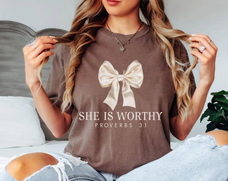 May include: A brown t-shirt with a white bow and the text "She is Worthy Proverbs 31" printed on the front.