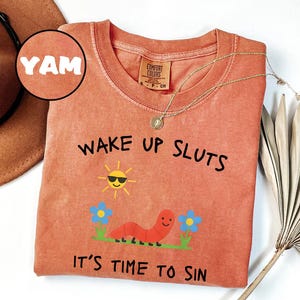 Puede incluir: Camiseta de color coral con el texto "WAKE UP SLUTS IT'S TIME TO SIN". El gr&aacute;fico incluye un sol sonriente con gafas de sol, un gusano rojo y flores azules. Un collar dorado est&aacute; drapeado sobre la camiseta.