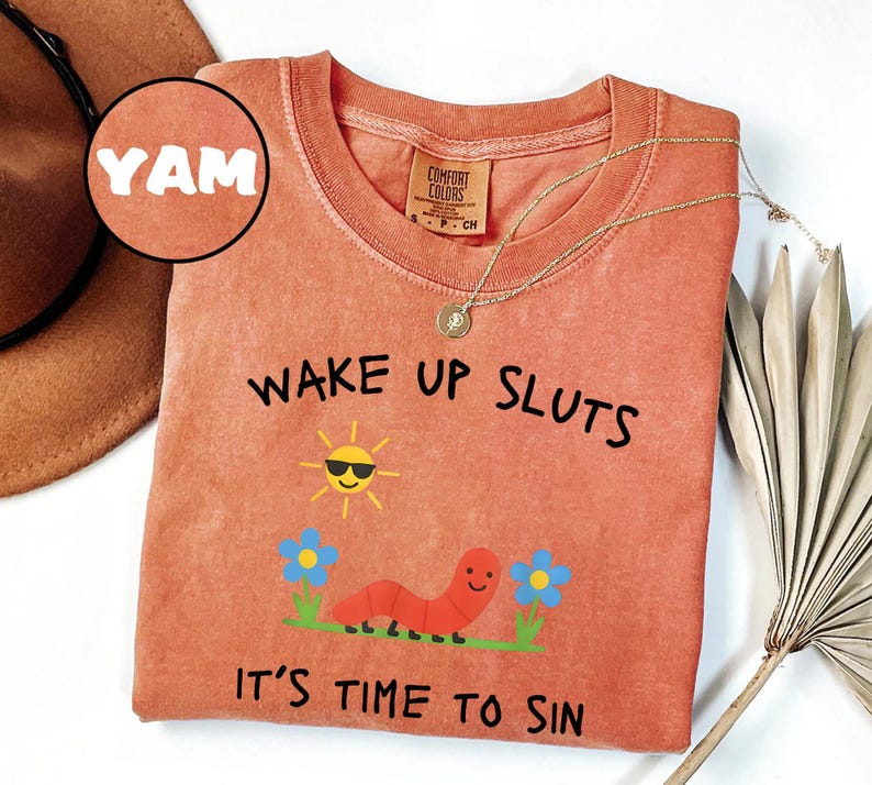 Puede incluir: Camiseta de color coral con el texto "WAKE UP SLUTS IT'S TIME TO SIN". El gr&aacute;fico incluye un sol sonriente con gafas de sol, un gusano rojo y flores azules. Un collar dorado est&aacute; drapeado sobre la camiseta.