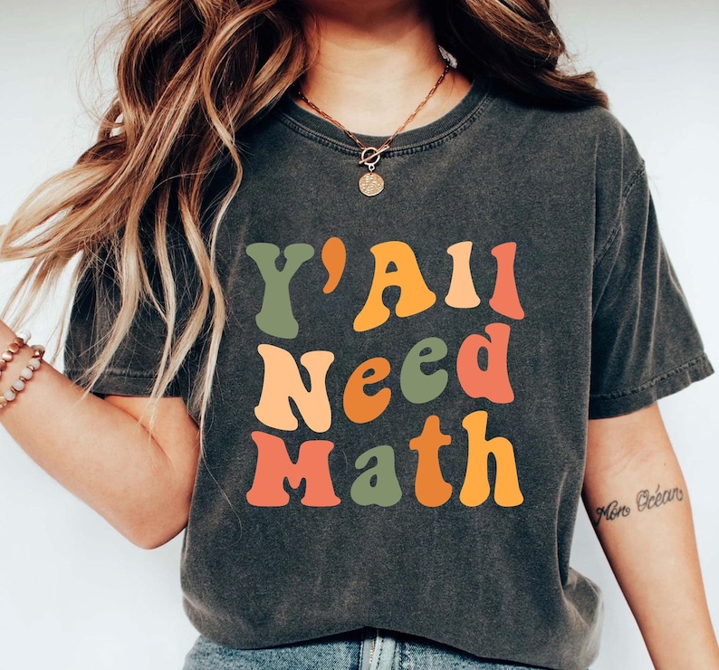 K&ouml;nnte beinhalten: Ein dunkelgraues T-Shirt mit dem Text "Y'all Need Math" in bunten, Retro-Lettern.