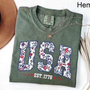 May include: Olive green t-shirt with the word "USA" in a floral design. The shirt also features the text "EST. 1776" in white. A silver necklace with a pendant is draped across the shirt. The shirt is folded on a white surface.