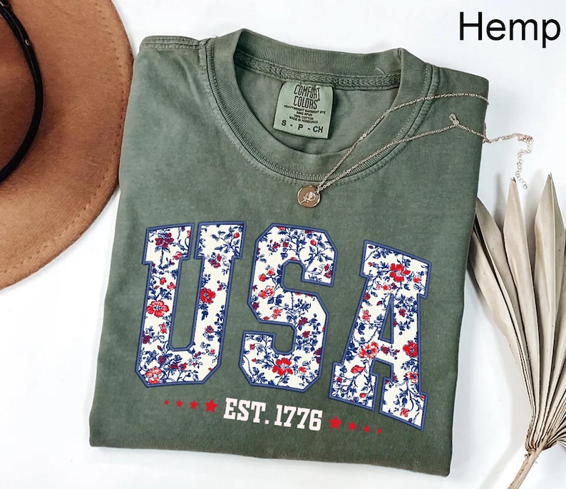 May include: Olive green t-shirt with the word "USA" in a floral design. The shirt also features the text "EST. 1776" in white. A silver necklace with a pendant is draped across the shirt. The shirt is folded on a white surface.