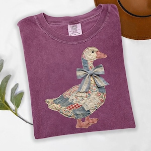 Peut inclure: T-shirt Comfort Colors violet avec un motif d'oie appliqu&eacute;. L'oie est faite de tissu patchwork avec des motifs floraux, vichy et unis. L'oie a un n&oelig;ud vichy bleu et des pieds vichy rouges. Le t-shirt a un col rond.