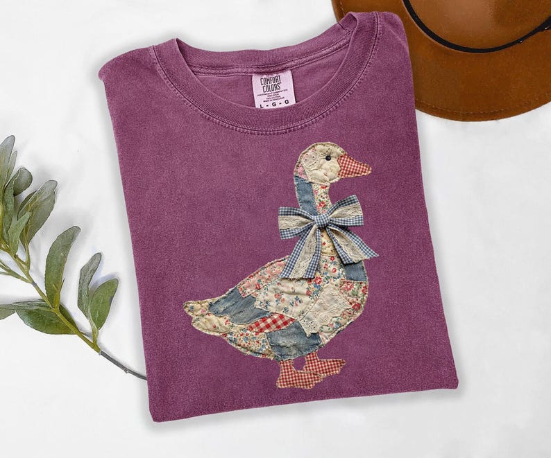 Peut inclure: T-shirt Comfort Colors violet avec un motif d'oie appliqu&eacute;. L'oie est faite de tissu patchwork avec des motifs floraux, vichy et unis. L'oie a un n&oelig;ud vichy bleu et des pieds vichy rouges. Le t-shirt a un col rond.
