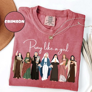 May include: Crimson-colored t-shirt with the phrase "Pray like a girl" in white script. The shirt features an illustration of seven figures in religious attire. A silver necklace with a pendant rests on the shirt.