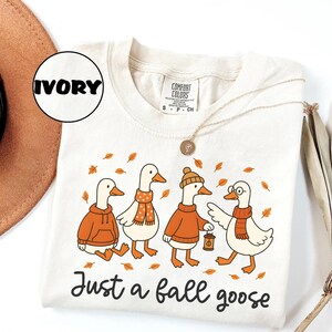 Puede incluir: Camiseta Comfort Colors color marfil con una ilustraci&oacute;n caprichosa de cuatro gansos vestidos con atuendos oto&ntilde;ales. Los gansos llevan su&eacute;teres, bufandas y un sombrero, con hojas de oto&ntilde;o esparcidas alrededor. El texto "Just a fall goose" est&aacute; impreso debajo.