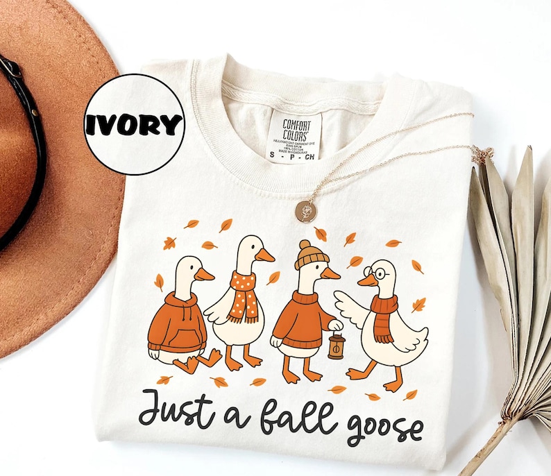 Puede incluir: Camiseta Comfort Colors color marfil con una ilustraci&oacute;n caprichosa de cuatro gansos vestidos con atuendos oto&ntilde;ales. Los gansos llevan su&eacute;teres, bufandas y un sombrero, con hojas de oto&ntilde;o esparcidas alrededor. El texto "Just a fall goose" est&aacute; impreso debajo.