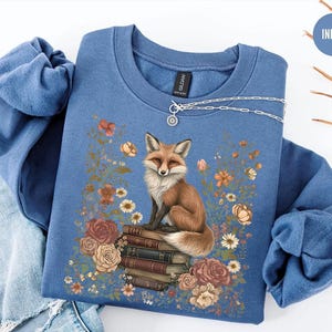 K&ouml;nnte beinhalten: Ein indigoblaues Sweatshirt mit Rundhalsausschnitt und einem Grafikdruck eines Fuchses, der auf einem Stapel B&uuml;cher sitzt, umgeben von Blumen in Orange-, Rosa- und Wei&szlig;t&ouml;nen. Eine silberne Halskette liegt auf dem Kragen des Sweatshirts.