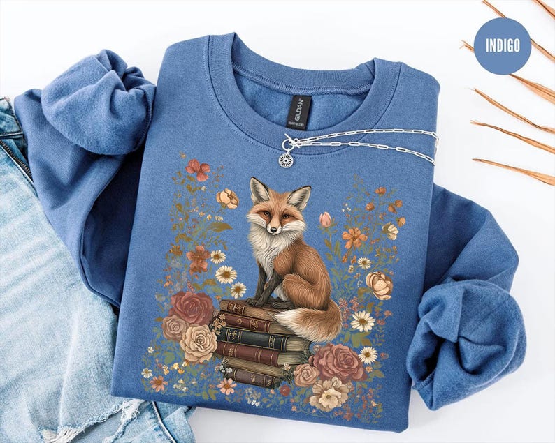 K&ouml;nnte beinhalten: Ein indigoblaues Sweatshirt mit Rundhalsausschnitt und einem Grafikdruck eines Fuchses, der auf einem Stapel B&uuml;cher sitzt, umgeben von Blumen in Orange-, Rosa- und Wei&szlig;t&ouml;nen. Eine silberne Halskette liegt auf dem Kragen des Sweatshirts.