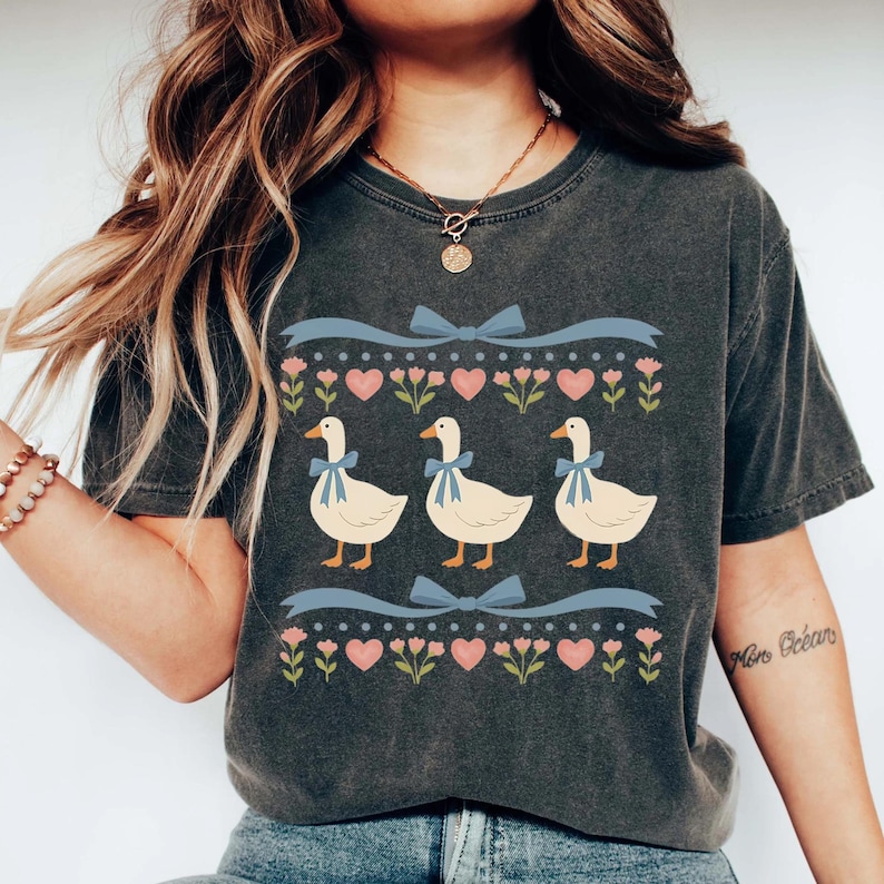 May include: Dark gray t-shirt featuring a whimsical design of three white geese with blue bows around their necks. The geese are set against a backdrop of pink hearts, flowers, and blue ribbons, creating a charming and playful aesthetic.