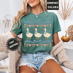 Pode incluir: Camiseta verde claro com um desenho de tr&ecirc;s gansos com la&ccedil;os azuis, cora&ccedil;&otilde;es e flores. A camiseta tem as palavras "COMFORT COLORS" e "LIGHT GREEN" impressas. &Eacute; uma pe&ccedil;a de roupa.