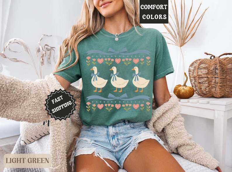 Pode incluir: Camiseta verde claro com um desenho de tr&ecirc;s gansos com la&ccedil;os azuis, cora&ccedil;&otilde;es e flores. A camiseta tem as palavras "COMFORT COLORS" e "LIGHT GREEN" impressas. &Eacute; uma pe&ccedil;a de roupa.