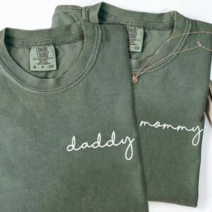Pu&ograve; includere: Due magliette a girocollo color verde oliva in cotone. Una maglietta presenta la scritta daddy in corsivo bianco, l'altra presenta la scritta mommy in corsivo bianco. Entrambe le magliette hanno un'etichetta del marchio Comfort Colors sul collo.