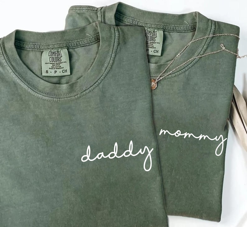 Pu&ograve; includere: Due magliette a girocollo color verde oliva in cotone. Una maglietta presenta la scritta daddy in corsivo bianco, l'altra presenta la scritta mommy in corsivo bianco. Entrambe le magliette hanno un'etichetta del marchio Comfort Colors sul collo.