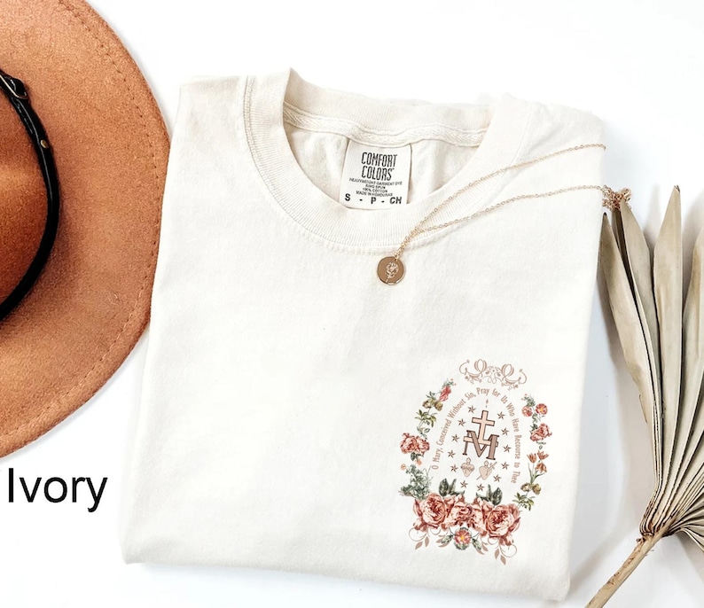 May include: Ivory t-shirt with a floral design featuring a Miraculous Medal. The design includes the words "Mary, Mother of Mercy, Pray for us now and at the hour of our death."