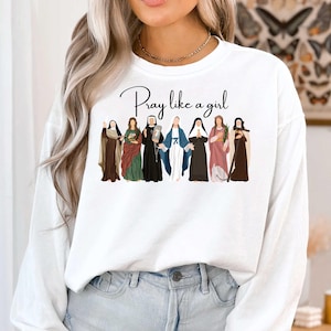 May include: White sweatshirt with the text "Pray like a girl" in black script. The design features illustrations of seven women in various outfits, including robes and veils, in shades of brown, green, black, blue, and pink.