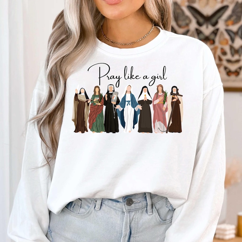 May include: White sweatshirt with the text "Pray like a girl" in black script. The design features illustrations of seven women in various outfits, including robes and veils, in shades of brown, green, black, blue, and pink.
