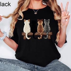 May include: Black crew neck t-shirt with a graphic of four cats on a wooden bench. The cats are white, black, brown, and grey. The word "Black" is printed in the upper left corner.