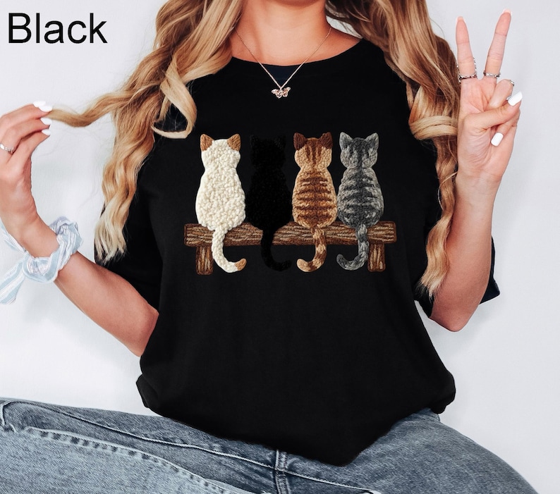May include: Black crew neck t-shirt with a graphic of four cats on a wooden bench. The cats are white, black, brown, and grey. The word "Black" is printed in the upper left corner.