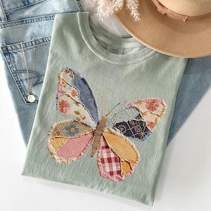 May include: A light green t-shirt with a patchwork butterfly design. The butterfly wings are made of various patterned fabrics in shades of blue, pink, yellow, and red. A pair of light blue jeans and a tan hat are also in the image.