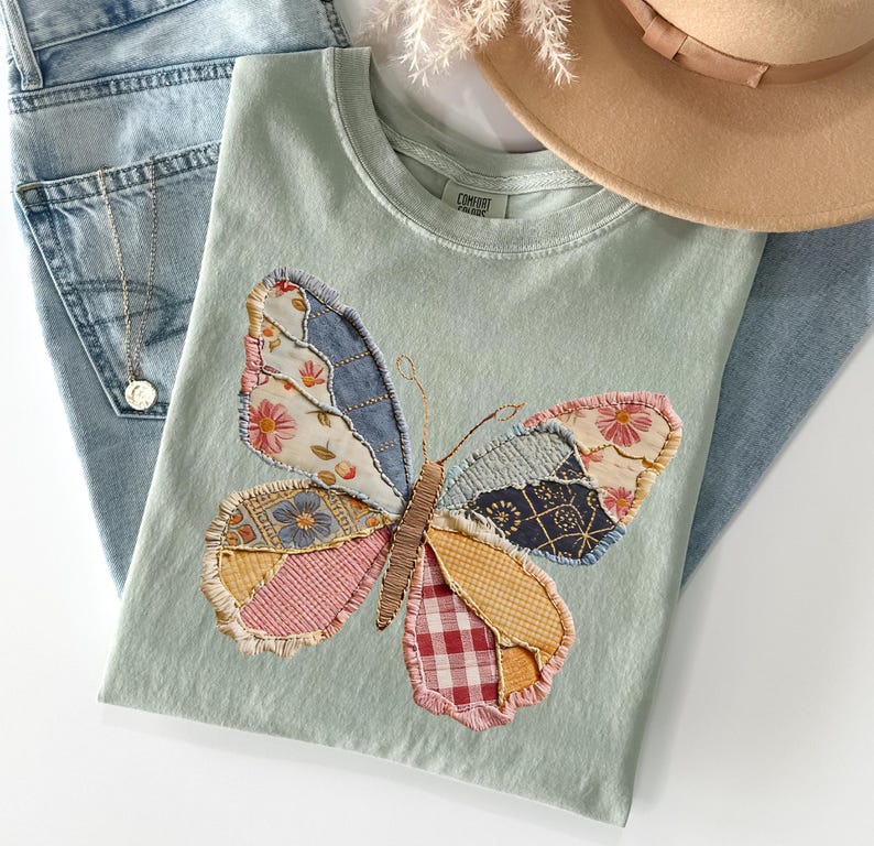 May include: A light green t-shirt with a patchwork butterfly design. The butterfly wings are made of various patterned fabrics in shades of blue, pink, yellow, and red. A pair of light blue jeans and a tan hat are also in the image.