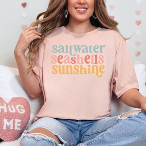 May include: A pale pink t-shirt featuring the words "saltwater seashells sunshine" in pastel shades. The shirt is worn with blue jeans. The design evokes a beach or summer theme, perfect for casual wear.