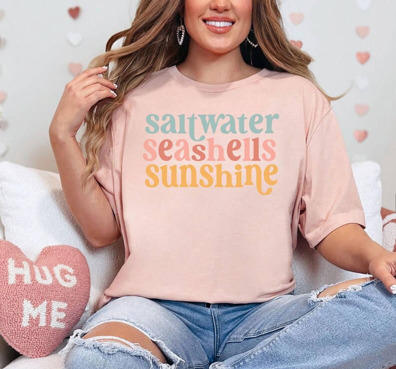 May include: A pale pink t-shirt featuring the words "saltwater seashells sunshine" in pastel shades. The shirt is worn with blue jeans. The design evokes a beach or summer theme, perfect for casual wear.