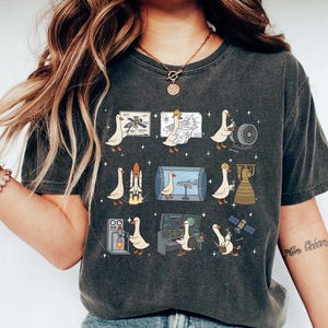May include: A charcoal gray short-sleeve t-shirt featuring a graphic print of nine white geese engaged in various aerospace engineering tasks, including working with rockets, satellites, blueprints, and control panels, surrounded by small white stars.