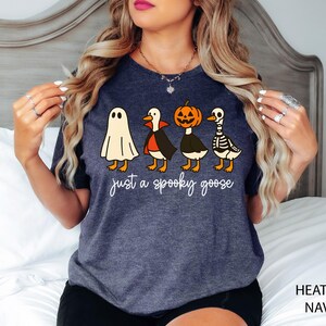 以下が含まれることがあります： ハロウィーンのデザインが施された、ヘザーネイビーのTシャツ。お化け、吸血鬼、カボチャ、骸骨に扮した4羽の漫画のガチョウが描かれ、白い筆記体で「just a spooky goose」と書かれています。楽しくてお祝いの服です。