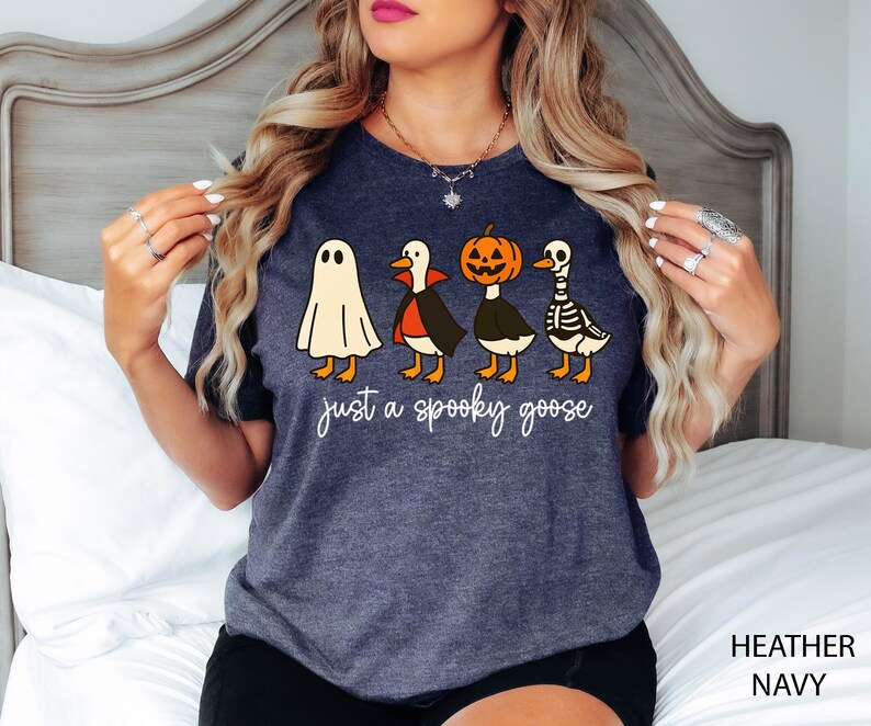 以下が含まれることがあります： ハロウィーンのデザインが施された、ヘザーネイビーのTシャツ。お化け、吸血鬼、カボチャ、骸骨に扮した4羽の漫画のガチョウが描かれ、白い筆記体で「just a spooky goose」と書かれています。楽しくてお祝いの服です。