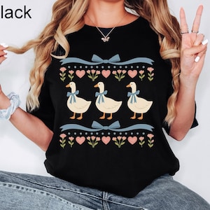 Pode incluir: Camiseta preta com um design l&uacute;dico com tr&ecirc;s gansos usando la&ccedil;os azuis, em um fundo de cora&ccedil;&otilde;es rosa, flores e fitas azuis. A palavra "Black" est&aacute; impressa no canto superior esquerdo.